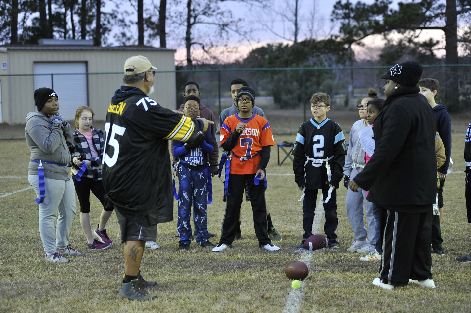 A coach giving instructions to a circle of young flag football players at dusk